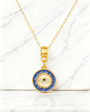 Colar Dourado Medalha Olho Grego Auvane em fundo de mármore claro com veios elegantes